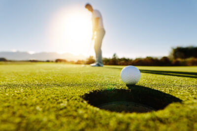 Pro golfer putting golf ball in to the hole. Golf ball by the hole with player in background on a sunny day.